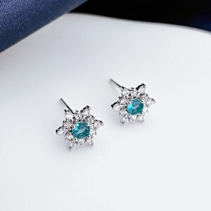 Op de afbeelding: Een paar zilveren oorstekers. Elke oorbel heeft een centrale turquoise edelsteen omringd door kleinere, heldere edelstenen, waardoor een sneeuwvlokpatroon ontstaat. De oorbellen zijn op een witte achtergrond.