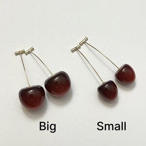 K&ouml;nnte beinhalten: Zwei Paar Kirschohrringe, eines mit der Aufschrift "Big" und das andere mit "Small". Die Kirschen sind tiefrot mit gl&auml;nzendem Finish, befestigt an goldfarbenen Stielen und Stiften. Die Ohrringe werden auf einer wei&szlig;en Oberfl&auml;che pr&auml;sentiert.