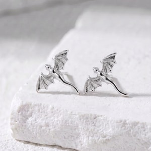 May include: A pair of silver-colored dragon-shaped stud earrings. Each earring features a detailed dragon with outstretched wings. The earrings are displayed on a white, textured surface, showcasing the intricate design and metallic sheen.