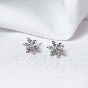 Op de afbeelding: Een paar zilveren sneeuwvlok oorstekers met heldere, sprankelende accenten. De oorbellen zijn geplaatst tegen een witte stoffen achtergrond, wat hun delicate ontwerp benadrukt. Een perfect accessoire voor de winter of feestdagen.