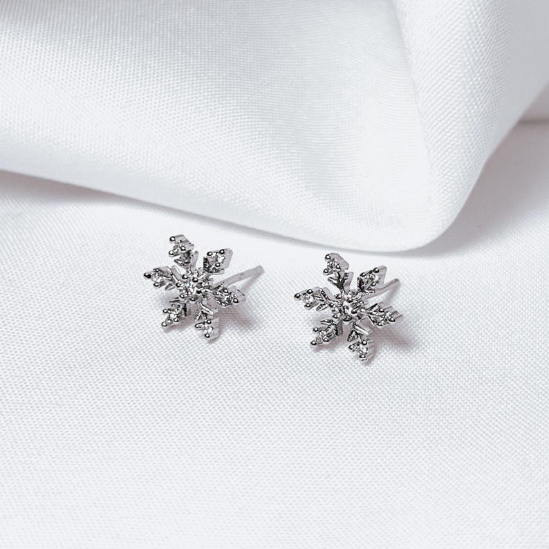 Op de afbeelding: Een paar zilveren sneeuwvlok oorstekers met heldere, sprankelende accenten. De oorbellen zijn geplaatst tegen een witte stoffen achtergrond, wat hun delicate ontwerp benadrukt. Een perfect accessoire voor de winter of feestdagen.