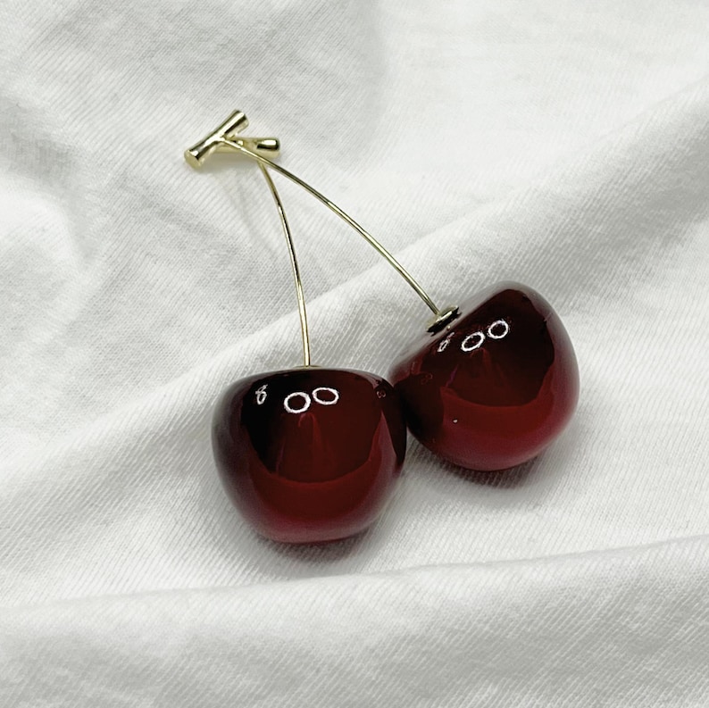 K&ouml;nnte beinhalten: Ein Paar Kirschohrringe mit goldfarbenem Stiel und goldfarbenem Verschluss. Die Kirschen sind tiefrot mit gl&auml;nzendem Finish und kleinen wei&szlig;en Akzenten. Die Ohrringe werden auf wei&szlig;em Stoffhintergrund pr&auml;sentiert.