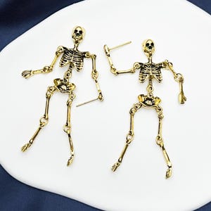 May include: A pair of gold-toned metal skeleton earrings featuring articulated limbs connected by small jump rings. Each earring depicts a full human skeleton figure with a detailed rib cage, skull, and jointed arms and legs, finished with a post back.