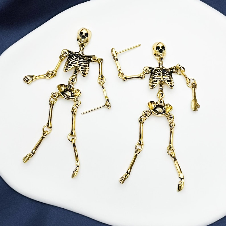 May include: A pair of gold-toned metal skeleton earrings featuring articulated limbs connected by small jump rings. Each earring depicts a full human skeleton figure with a detailed rib cage, skull, and jointed arms and legs, finished with a post back.