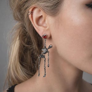 May include: A silver-toned skeleton dangle earring featuring a small, faceted red heart-shaped gemstone at the top post. The articulated skeleton figure hangs from the heart, with movable limbs and a detailed skull, torso, and pelvic structure.