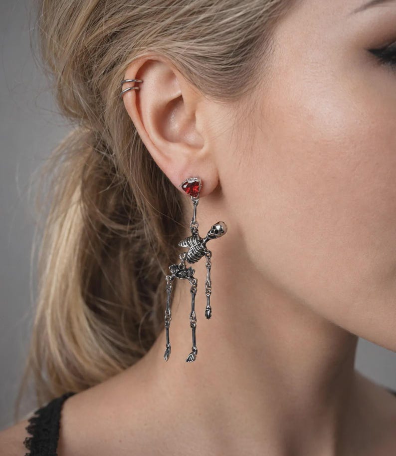May include: A silver-toned skeleton dangle earring featuring a small, faceted red heart-shaped gemstone at the top post. The articulated skeleton figure hangs from the heart, with movable limbs and a detailed skull, torso, and pelvic structure.