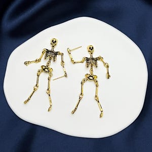 May include: A pair of gold-toned skeleton earrings featuring articulated limbs and a detailed rib cage design. Each earring is constructed with multiple connected segments that allow for movement, finished with a post back for secure attachment.