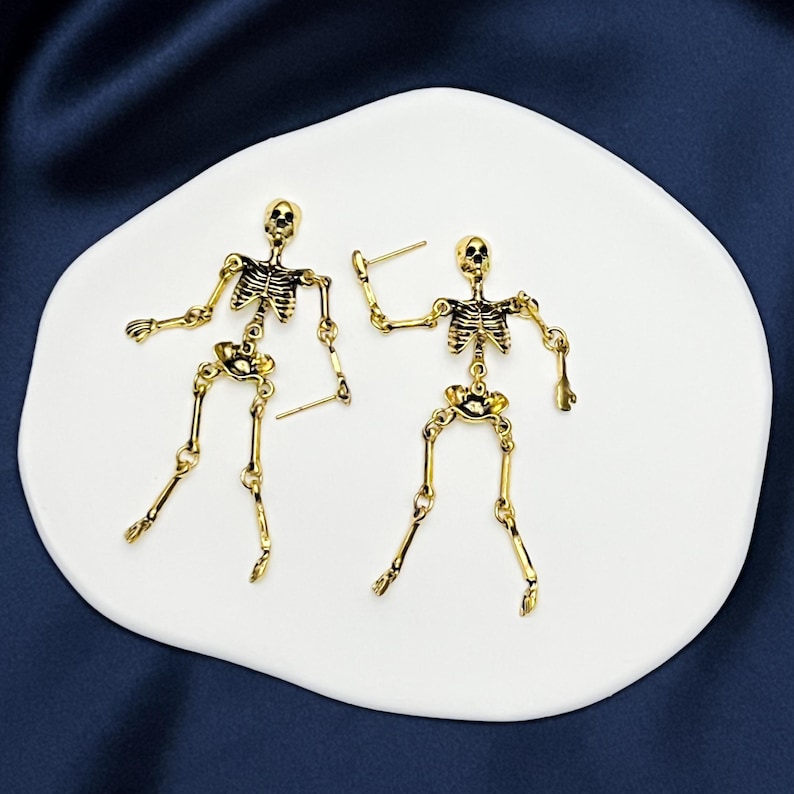 May include: A pair of gold-toned skeleton earrings featuring articulated limbs and a detailed rib cage design. Each earring is constructed with multiple connected segments that allow for movement, finished with a post back for secure attachment.