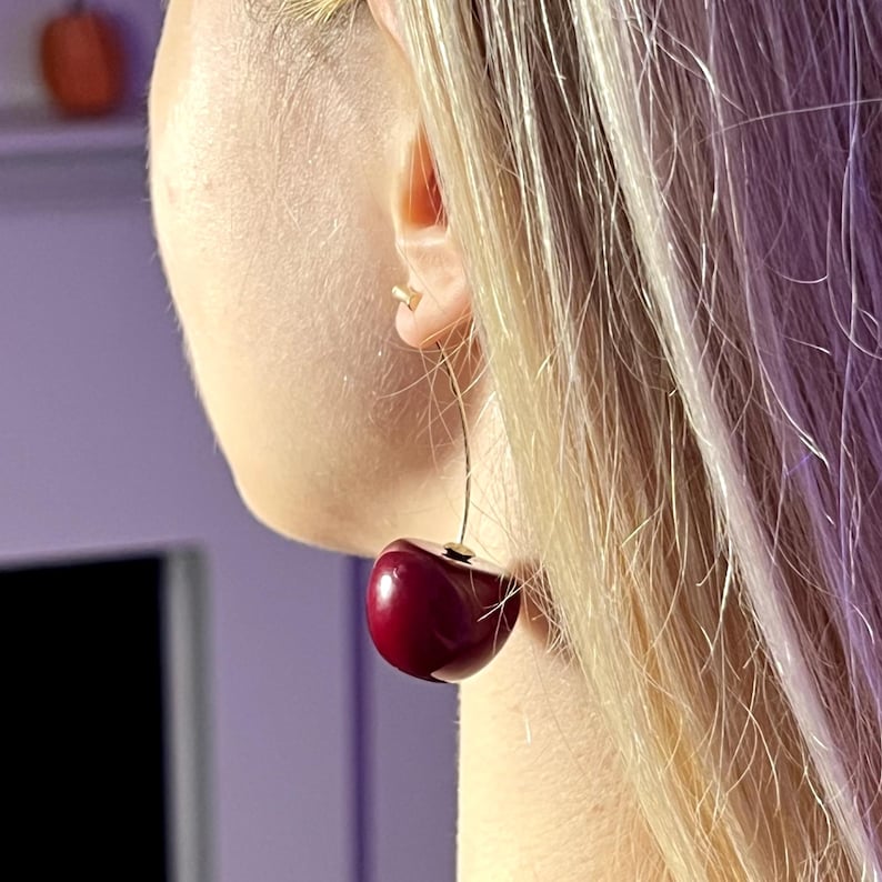 K&ouml;nnte beinhalten: Nahaufnahme eines Kirschohrrings. Der Ohrring zeigt eine gro&szlig;e, gl&auml;nzende, dunkelrote Kirsche, die an einem d&uuml;nnen, goldfarbenen Draht h&auml;ngt. Der Ohrring wird am Ohr einer Person mit blondem Haar getragen.