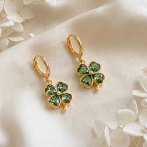 Peut inclure: Une paire de boucles d'oreilles cr&eacute;oles dor&eacute;es avec des pendentifs en forme de tr&egrave;fle &agrave; quatre feuilles. Chaque tr&egrave;fle est compos&eacute; de quatre p&eacute;tales en cristal vert fonc&eacute; taill&eacute;s en forme de c&oelig;ur, sertis dans une monture en m&eacute;tal dor&eacute;.