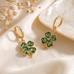 Peut inclure: Une paire de boucles d'oreilles cr&eacute;oles dor&eacute;es avec des pendentifs en forme de tr&egrave;fle &agrave; quatre feuilles. Chaque tr&egrave;fle est compos&eacute; de quatre cristaux vert fonc&eacute; facett&eacute;s sertis dans une monture en m&eacute;tal dor&eacute;, suspendus &agrave; une cr&eacute;ole simple.