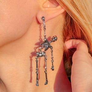 May include: A silver-toned dangling earring shaped like a human skeleton. The skeleton figure is articulated with separate pieces for the torso, pelvis, and legs, connected by small metal rings, hanging from a post that attaches at the skull.