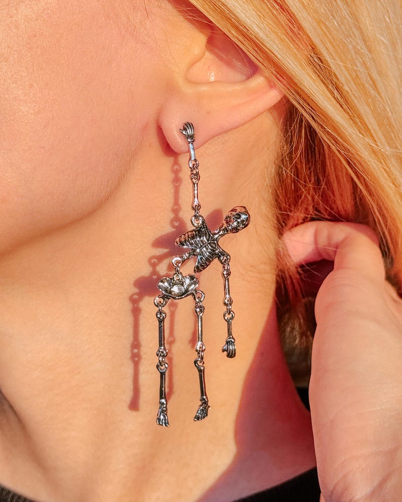 May include: A silver-toned dangling earring shaped like a human skeleton. The skeleton figure is articulated with separate pieces for the torso, pelvis, and legs, connected by small metal rings, hanging from a post that attaches at the skull.