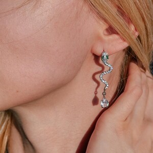 Peut inclure: Boucle d'oreille argentée en forme de serpent, ornée d'une gemme verte à la tête et de gemmes claires le long du corps. Une gemme claire en forme de goutte pend du bas du serpent.