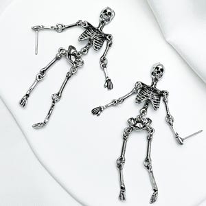 May include: A pair of silver-toned metal earrings shaped like articulated human skeletons. Each earring features a detailed skull, rib cage, pelvic bone, and jointed limbs connected by small metal rings, with a post back for pierced ears.
