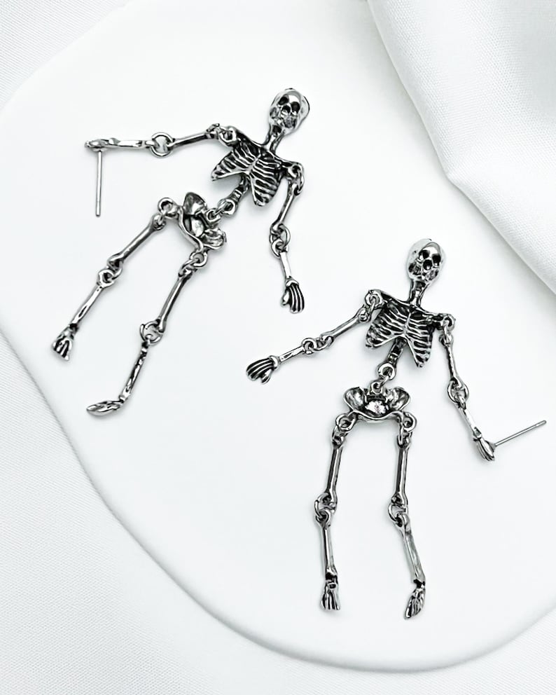 May include: A pair of silver-toned metal earrings shaped like articulated human skeletons. Each earring features a detailed skull, rib cage, pelvic bone, and jointed limbs connected by small metal rings, with a post back for pierced ears.
