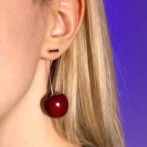 K&ouml;nnte beinhalten: Nahaufnahme eines Ohrs mit einem Kirschohrring. Der Ohrring hat einen gro&szlig;en, gl&auml;nzenden, dunkelroten Kirschenanh&auml;nger, der an einem d&uuml;nnen, goldfarbenen Draht h&auml;ngt. Ein kleiner, schwarz-goldener Ohrstecker ist ebenfalls zu sehen.