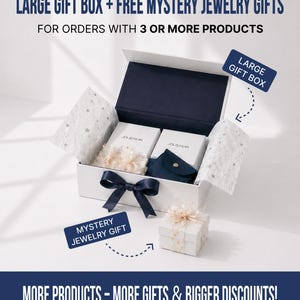 May include: A white rectangular gift box with a dark blue ribbon, containing two white boxes labeled JOLIS NOR, a dark blue pouch, and star-patterned tissue paper. A smaller white gift box sits in front with text labels Large Gift Box and Mystery Jewelry Gift.