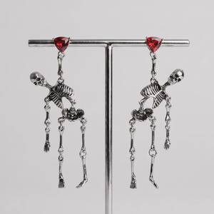 May include: A pair of silver-toned dangle earrings featuring articulated skeleton figures. Each earring is topped with a triangular, faceted red gemstone stud. The skeletons are depicted in a hanging pose with movable joints, crafted from polished metal with detailed bone structures.
