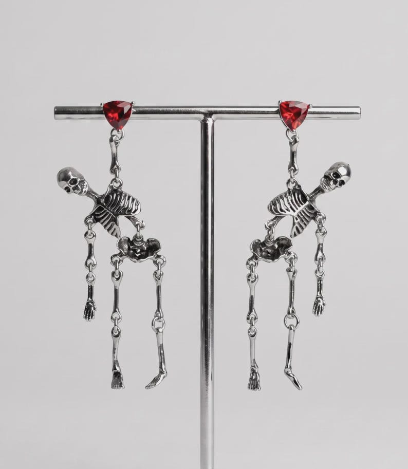 May include: A pair of silver-toned dangle earrings featuring articulated skeleton figures. Each earring is topped with a triangular, faceted red gemstone stud. The skeletons are depicted in a hanging pose with movable joints, crafted from polished metal with detailed bone structures.