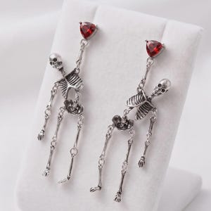 May include: A pair of silver-toned dangle earrings featuring articulated skeleton figures. Each earring is topped with a triangular, faceted red gemstone. The skeletons are constructed with linked metal segments, allowing for movement at the joints.