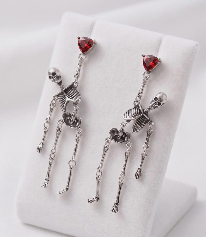 May include: A pair of silver-toned dangle earrings featuring articulated skeleton figures. Each earring is topped with a triangular, faceted red gemstone. The skeletons are constructed with linked metal segments, allowing for movement at the joints.