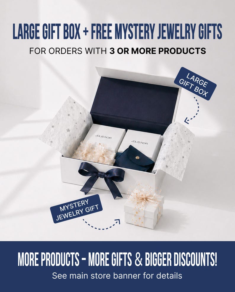 Op de afbeelding: Een witte rechthoekige geschenkdoos met een donkerblauw lint, met daarin twee witte doosjes met het label JOLIS NOR en een donkerblauw zakje. De tekst luidt: LARGE GIFT BOX + FREE MYSTERY JEWELRY GIFTS FOR ORDERS WITH 3 OR MORE PRODUCTS en MORE PRODUCTS - MORE GIFTS & BIGGER DISCOUNTS!