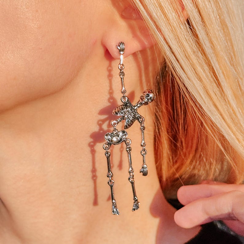 May include: A silver-toned dangle earring shaped like a full human skeleton. The articulated figure features a detailed skull, rib cage, and limbs that hang vertically from a small hook. The metal has a polished finish with dark recessed areas.