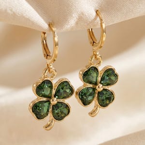 Peut inclure: Une paire de boucles d'oreilles cr&eacute;oles dor&eacute;es avec des pendentifs en forme de tr&egrave;fle &agrave; quatre feuilles. Chaque tr&egrave;fle est compos&eacute; de quatre cristaux facett&eacute;s vert fonc&eacute; sertis dans une monture en m&eacute;tal dor&eacute;, suspendus &agrave; un petit anneau.