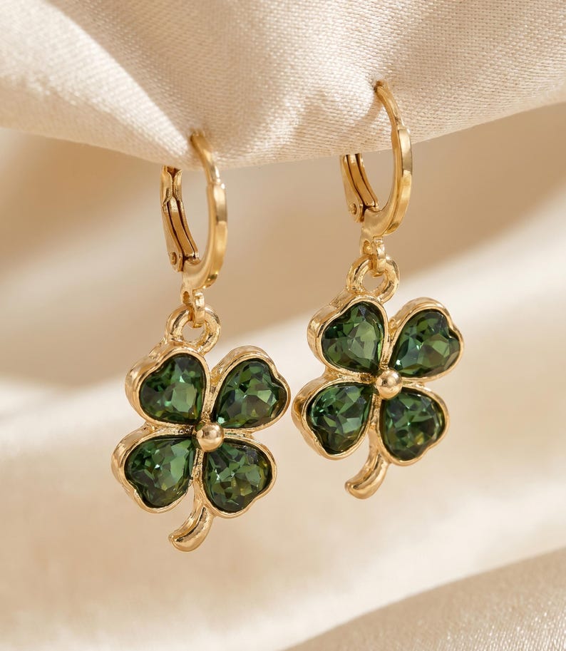 Peut inclure: Une paire de boucles d'oreilles cr&eacute;oles dor&eacute;es avec des pendentifs en forme de tr&egrave;fle &agrave; quatre feuilles. Chaque tr&egrave;fle est compos&eacute; de quatre cristaux facett&eacute;s vert fonc&eacute; sertis dans une monture en m&eacute;tal dor&eacute;, suspendus &agrave; un petit anneau.