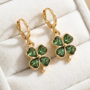 Peut inclure: Une paire de boucles d'oreilles cr&eacute;oles dor&eacute;es avec des pendentifs en forme de tr&egrave;fle &agrave; quatre feuilles. Chaque tr&egrave;fle est compos&eacute; de quatre cristaux verts facett&eacute;s sertis dans une monture en m&eacute;tal dor&eacute;, suspendus &agrave; une attache articul&eacute;e.