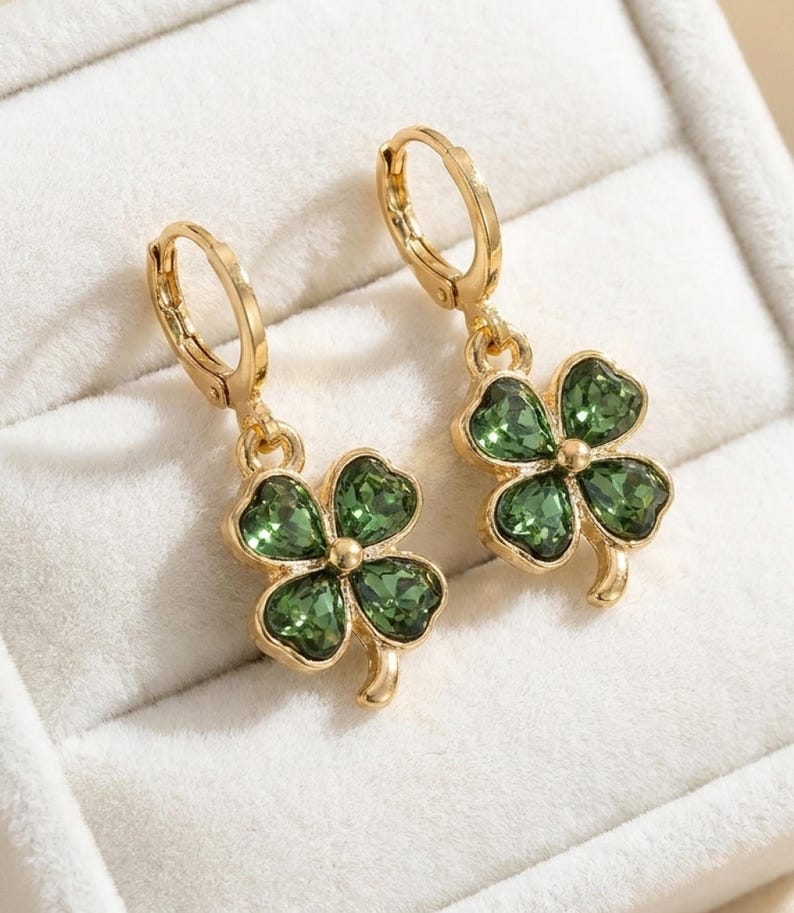 Peut inclure: Une paire de boucles d'oreilles cr&eacute;oles dor&eacute;es avec des pendentifs en forme de tr&egrave;fle &agrave; quatre feuilles. Chaque tr&egrave;fle est compos&eacute; de quatre cristaux verts facett&eacute;s sertis dans une monture en m&eacute;tal dor&eacute;, suspendus &agrave; une attache articul&eacute;e.