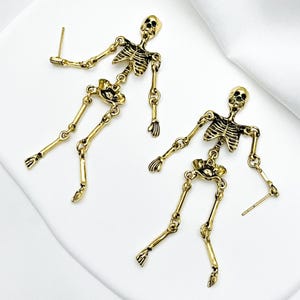 May include: A pair of gold-toned metal skeleton earrings featuring articulated limbs and a detailed rib cage design. Each earring has a post back closure and depicts a full human skeletal figure with a skull, torso, arms, and legs in a dangling style.