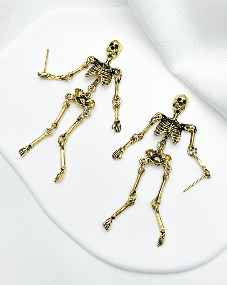 May include: A pair of gold-toned metal skeleton earrings featuring articulated limbs and a detailed rib cage design. Each earring has a post back closure and depicts a full human skeletal figure with a skull, torso, arms, and legs in a dangling style.
