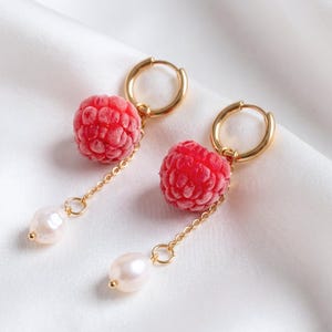 May include: A pair of gold hoop earrings featuring a textured red raspberry charm and a dangling white freshwater pearl. Each earring consists of a circular gold hoop, a realistic red berry pendant, and a delicate gold chain ending in a pearl.
