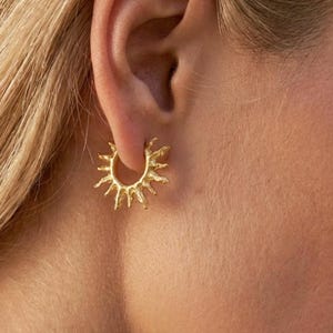 Op de afbeelding: Een gouden oorring met een getextureerd, op de zon ge&iuml;nspireerd ontwerp met stralende punten die uitsteken vanaf de cirkelvormige band. Het metalen oppervlak heeft een matte, organische afwerking en zit vast in de oorlel.