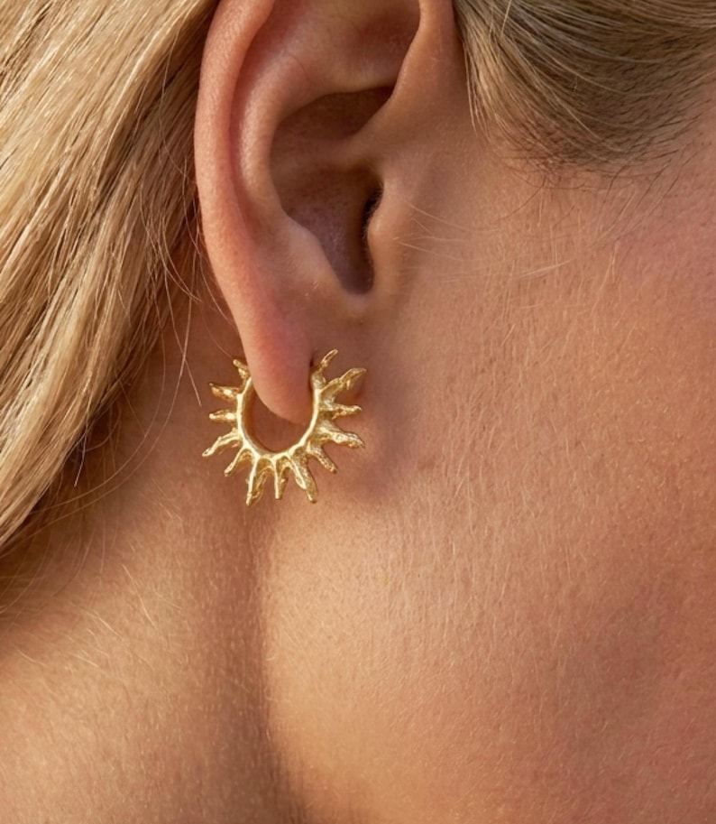 Op de afbeelding: Een gouden oorring met een getextureerd, op de zon ge&iuml;nspireerd ontwerp met stralende punten die uitsteken vanaf de cirkelvormige band. Het metalen oppervlak heeft een matte, organische afwerking en zit vast in de oorlel.