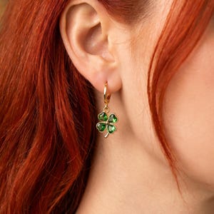 Peut inclure: Une boucle d'oreille cr&eacute;ole dor&eacute;e avec une breloque en forme de tr&egrave;fle &agrave; quatre feuilles. Le tr&egrave;fle est compos&eacute; de quatre pierres vertes translucides en forme de c&oelig;ur, serties dans une monture en m&eacute;tal dor&eacute;, suspendue &agrave; un anneau dor&eacute;.