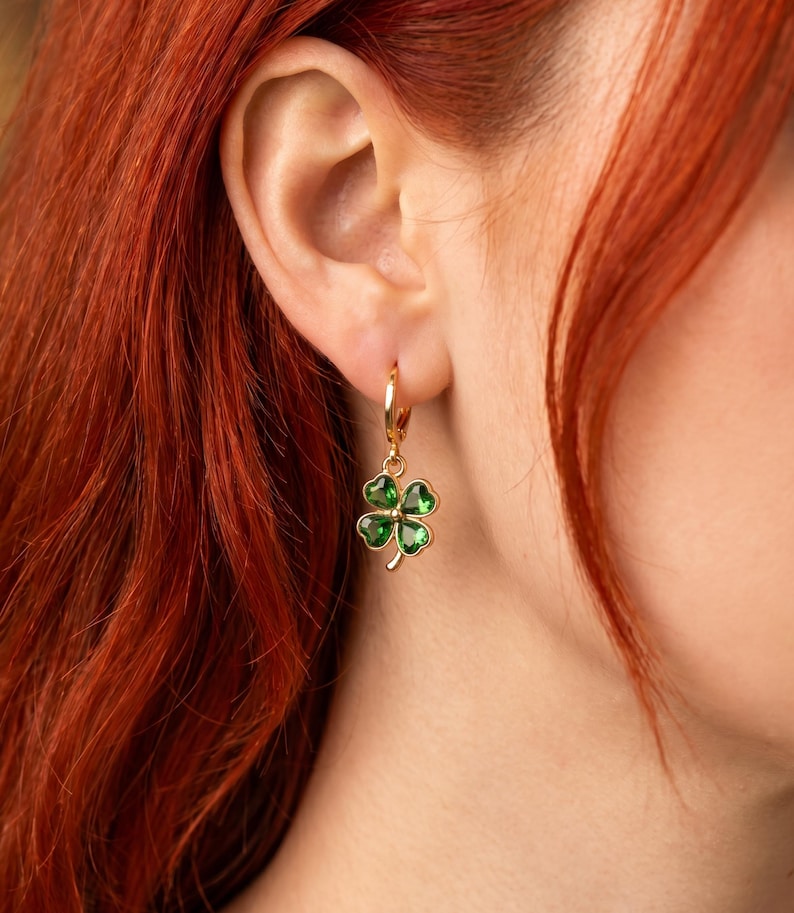 Peut inclure: Une boucle d'oreille cr&eacute;ole dor&eacute;e avec une breloque en forme de tr&egrave;fle &agrave; quatre feuilles. Le tr&egrave;fle est compos&eacute; de quatre pierres vertes translucides en forme de c&oelig;ur, serties dans une monture en m&eacute;tal dor&eacute;, suspendue &agrave; un anneau dor&eacute;.