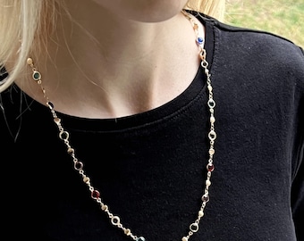May include: A gold-toned chain necklace featuring a series of small, round, multi-colored gemstones set at intervals along the length of the chain. The stones include various hues such as red, blue, green, and clear, connected by thin gold links.