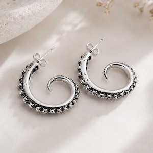 May include: A pair of silver hoop earrings shaped like octopus tentacles, featuring a curved design with raised circular suction cup details along the outer edge. Each earring includes a post back closure and is stamped with the number 923.