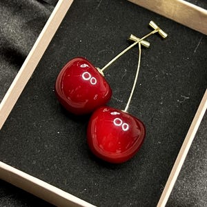 K&ouml;nnte beinhalten: Kirschohrringe. Jeder Ohrring hat eine gl&auml;nzende, tiefrote Kirsche mit einem goldfarbenen Stiel und einem T-f&ouml;rmigen Verschluss. Die Ohrringe werden in einer hellen Schachtel mit schwarzem Innenraum pr&auml;sentiert.