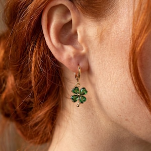 Peut inclure: Une boucle d'oreille cr&eacute;ole en or avec un pendentif en forme de tr&egrave;fle &agrave; quatre feuilles, compos&eacute; de quatre pierres vert &eacute;meraude facett&eacute;es serties dans une monture dor&eacute;e. Le tr&egrave;fle est suspendu &agrave; un petit anneau en or poli fix&eacute; au lobe.