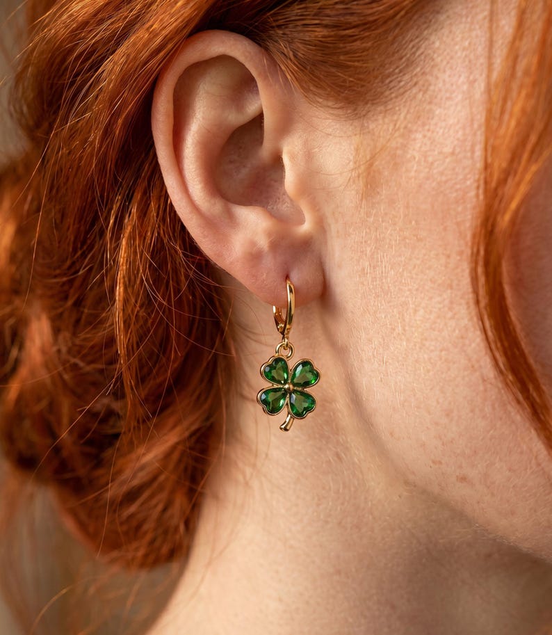 Peut inclure: Une boucle d'oreille cr&eacute;ole en or avec un pendentif en forme de tr&egrave;fle &agrave; quatre feuilles, compos&eacute; de quatre pierres vert &eacute;meraude facett&eacute;es serties dans une monture dor&eacute;e. Le tr&egrave;fle est suspendu &agrave; un petit anneau en or poli fix&eacute; au lobe.