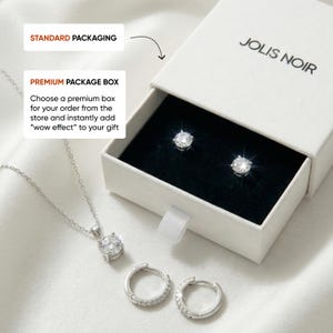 May include: A silver-toned jewelry set featuring a pendant necklace with a round clear stone, a pair of small hoop earrings, and two stud earrings with round clear stones. The items are displayed with a white JOLIS NOIR branded gift box.
