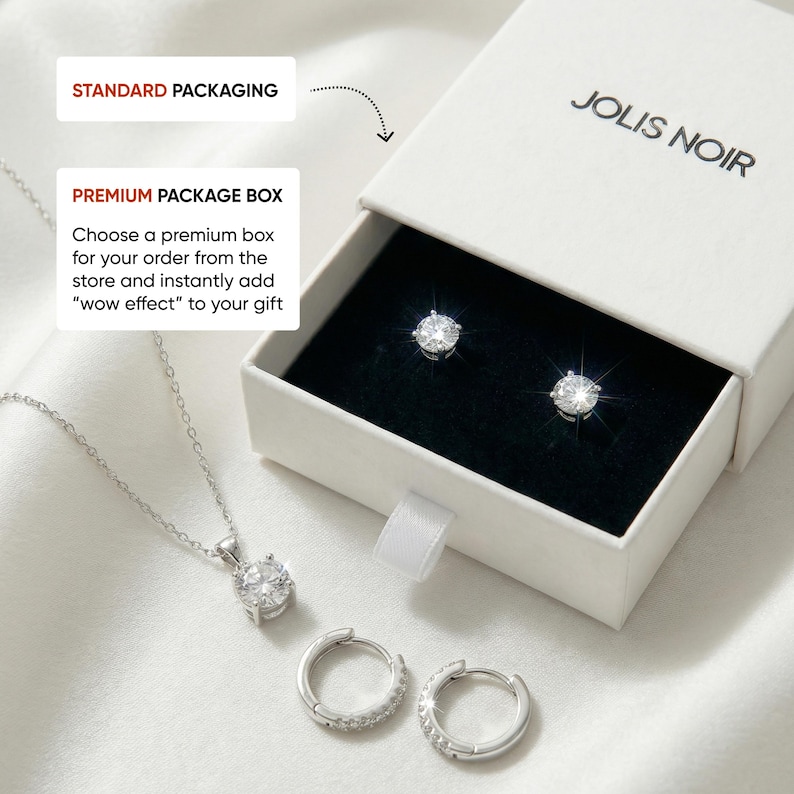 May include: A silver-toned jewelry set featuring a pendant necklace with a round clear stone, a pair of small hoop earrings, and two stud earrings with round clear stones. The items are displayed with a white JOLIS NOIR branded gift box.
