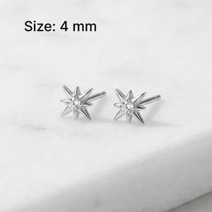 May include: A pair of silver-colored starburst stud earrings with a central clear stone. The earrings are 4 mm in size and are displayed on a white surface. The text "Size: 4 mm" is above the earrings.