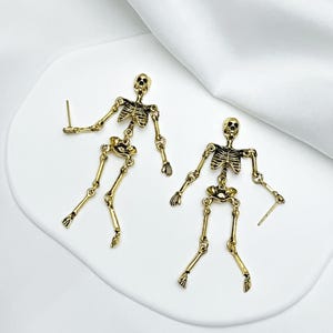May include: A pair of gold-toned metal skeleton earrings featuring articulated limbs and a detailed rib cage design. Each skeleton figure is suspended from a post backing, showcasing a metallic finish with darkened recessed areas to highlight the bone structure.
