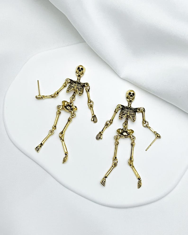 May include: A pair of gold-toned metal skeleton earrings featuring articulated limbs and a detailed rib cage design. Each skeleton figure is suspended from a post backing, showcasing a metallic finish with darkened recessed areas to highlight the bone structure.