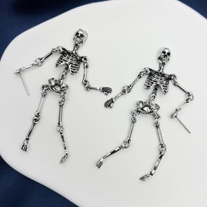 May include: A pair of silver-toned metal earrings shaped like articulated human skeletons. Each earring features a detailed skull, rib cage, pelvic bone, and jointed limbs connected by small metal rings, with a post backing for attachment.